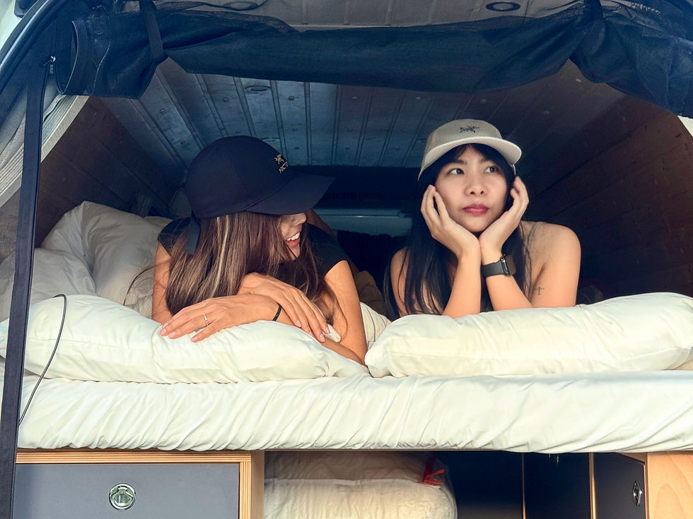 Two women lie on a bed in a van interior. One smiles, the other looks pensive. Both wear hats. White pillows and wood paneling visible. Japan campervan, Tokyo campervan, Tokyo, vanlife, Japan road trip, camper van