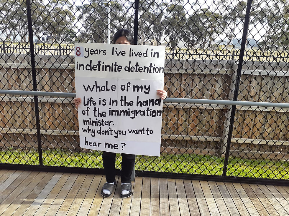 Woman stands in front of high fence, holding a sign that reads, '8 year I've lived in indefinite detention. Whole of my life is in the hands of the immigration minister. Why don't you want to hear me?'