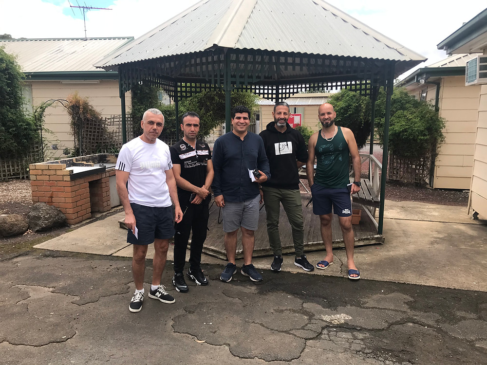 Five men standing in a line smiling at the camera with a bbq and patio in the background.