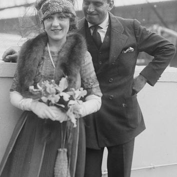 Elegant woman in fur hat holds flowers, man in suit smiles behind her.