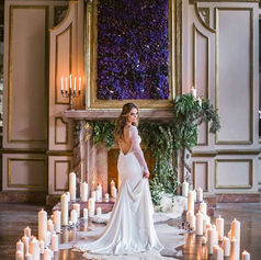 Woman in white wedding dress stands amidst many lit candles in ornate room.