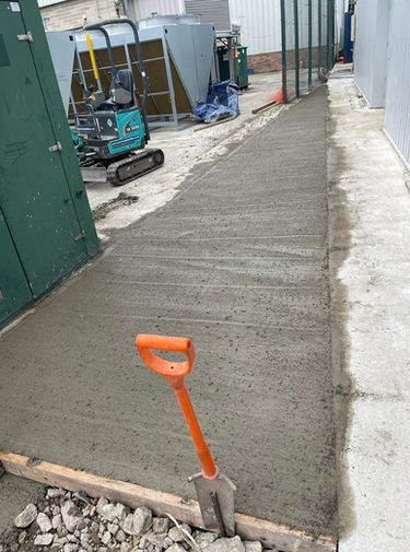 In an active construction site, a mini excavator sits idle on the rough concrete ground. A blue bag lies nearby, resting on the ground. An assortment of tools and equipment is scattered throughout the area, including a shovel leaning against a concrete wall and another shovel being used to dig a hole in the ground. A man pours concrete into a trench with a shovel, while another rides a skateboard on the concrete floor. The scene is dominated by a large building with green doors on its side and an excavator parked in front of it.