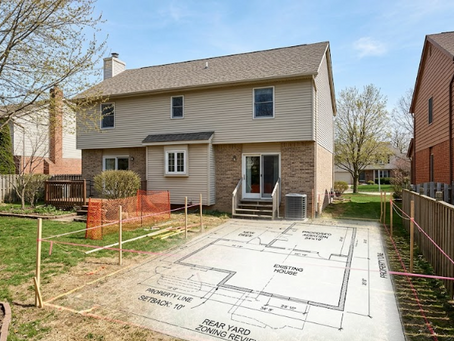 A high-angle photo of a suburban backyard in Northeast Ohio during early spring, featuring a two-story beige siding and brick home. The lawn is marked with pink surveyor's string and wooden stakes, with a blueprint architectural drawing of a proposed addition overlaid on the grass, highlighting proximity to neighboring fences and property lines under a clear blue sky.