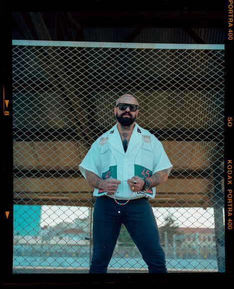 Bald man with beard, sunglasses, white shirt, poses confidently urban background.
