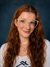 Sarah, Marketing Strategist at MPS Contracting, smiling in a white MPS shirt with long wavy auburn hair, glasses, and a blue studio background.