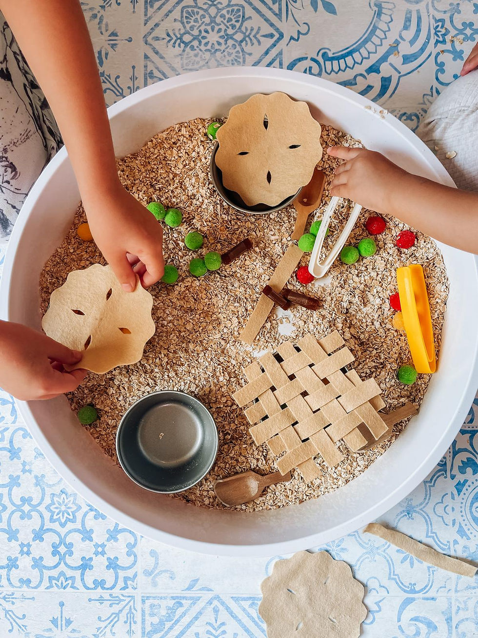 Children's hands playing with a, apple pie inspired sensory bin of oats, felt pieces, and toys. The bin is on a blue patterned floor, evoking playful creativity.