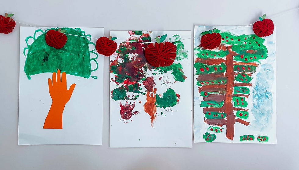 Three children's paintings with an apple tree theme, featuring handprints as leaves and trunk. Red apple garlands handmade of cardboard and string hang above on a white wall.
