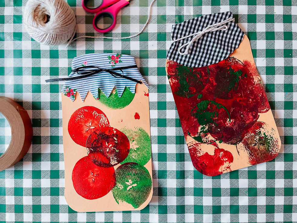 Crafted jar-shaped cards with red and green paint stamped with cut apples, on a green plaid tablecloth. Nearby, pink scissors and twine evoke a creative vibe.