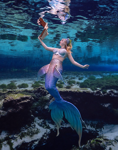 Mermaid Kalynn is reading a book underwater. She holds it up in front of her, sun skimming over the pages and her hair, sparkling off her bejeweled crown and across her blue scales.