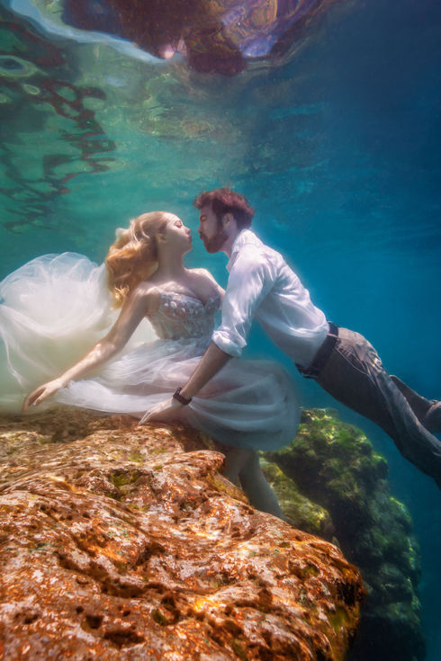 Kalynn is wearing a wedding dress underwater. She leans against a rock as her husband, Cody, swims in towards her. He wears khaki pants and a white button-down.