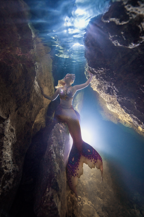 Mermaid Kalynn sits on a rock ledge underwater in a chasm of a cave. Bright light shines in the distance behind her as she stares into the dark waters. Her golden hair flows behind her as light beams shine down across her face.