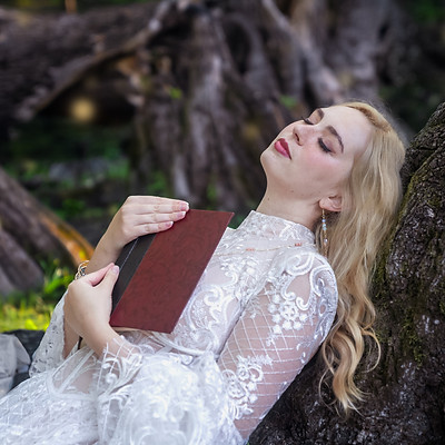Kalynn lays against a tree in a white lace dress. She appears to have fallen asleep reading, a book sits open on her chest. The forest around her is lush and green and practically sparkling with life.