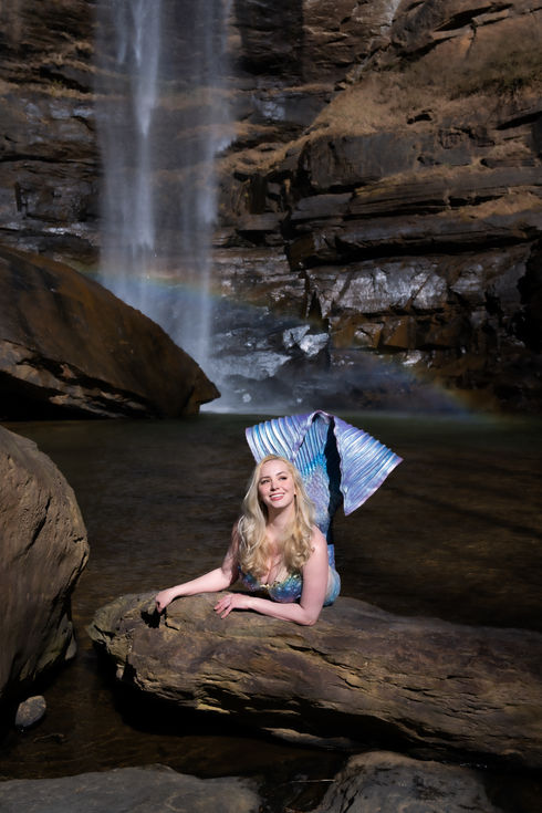 Mermaid Kalynn poses on the rocks in front of a waterfall. A rainbow appears in the mist coming off the pool, and she smiles.