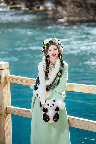 色鮮やかなチベット民族衣装に身を包んだ女性が、パンダのぬいぐるみを手に微笑む。背後にはエメラルドブルーの湖と山々が広がり、九寨溝の幻想的な美景が映し出されています。