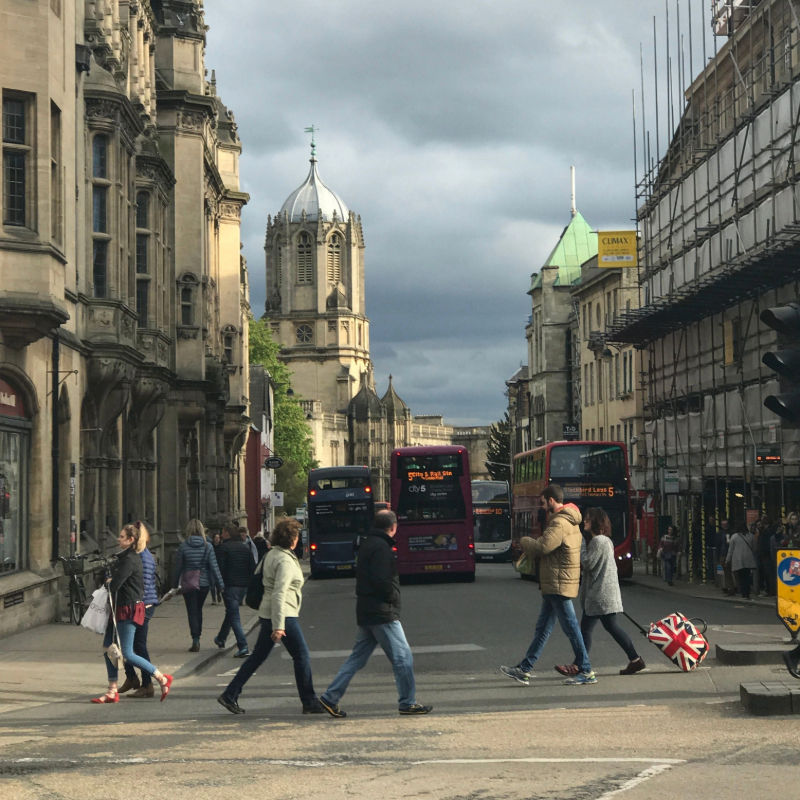 Bust street in Oxford with pedestrians crossing the road, double-decker buses in traffic and the domed Tom Tower visible in the background.