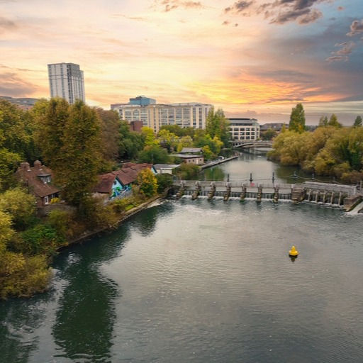 View of the river with a weir and lock structure in Reading, framed by leafy trees on the banks, riverside buildings, and high-rise structures with a sunset sky.
