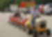 A barrel train! A man in a grey shirt driving a lawn mower that is towing 5 train cars made from barrels. Two barrel cars are accompanied, one by a small child wearing light blue ear muffs and one by a female adult. They are diving in a parking lot where there are two firetrucks in the background being viewed by other people at the event. One bystander is a female in a powered wheelchair.