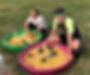 Two young adults playing in two different plastic pools that are filled with corn kernels and toy vehicles. The person on the left is a young lady that has down syndrome and the person on the right is a young man wearing a hulk mask that appears to also have down syndrome as well. The pair look like they are having so much fun, both with big smiles on their faces.