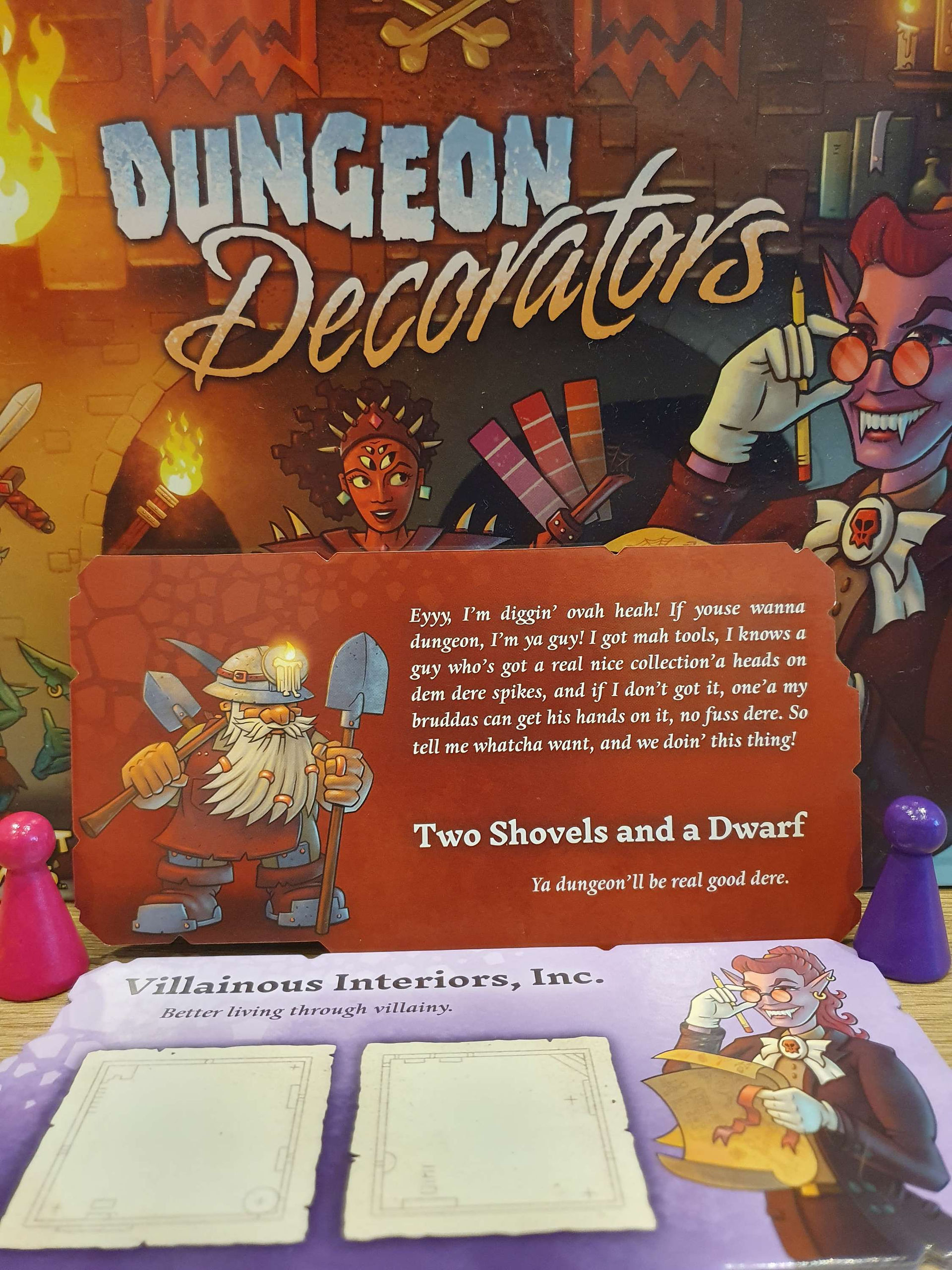 The 'Two Shovels and a Dwarf' design company card from the game Dungeon Decorators. It is a red card with a cartoon dwarf wearing a helmet and wielding two shovels and a pickaxe. 