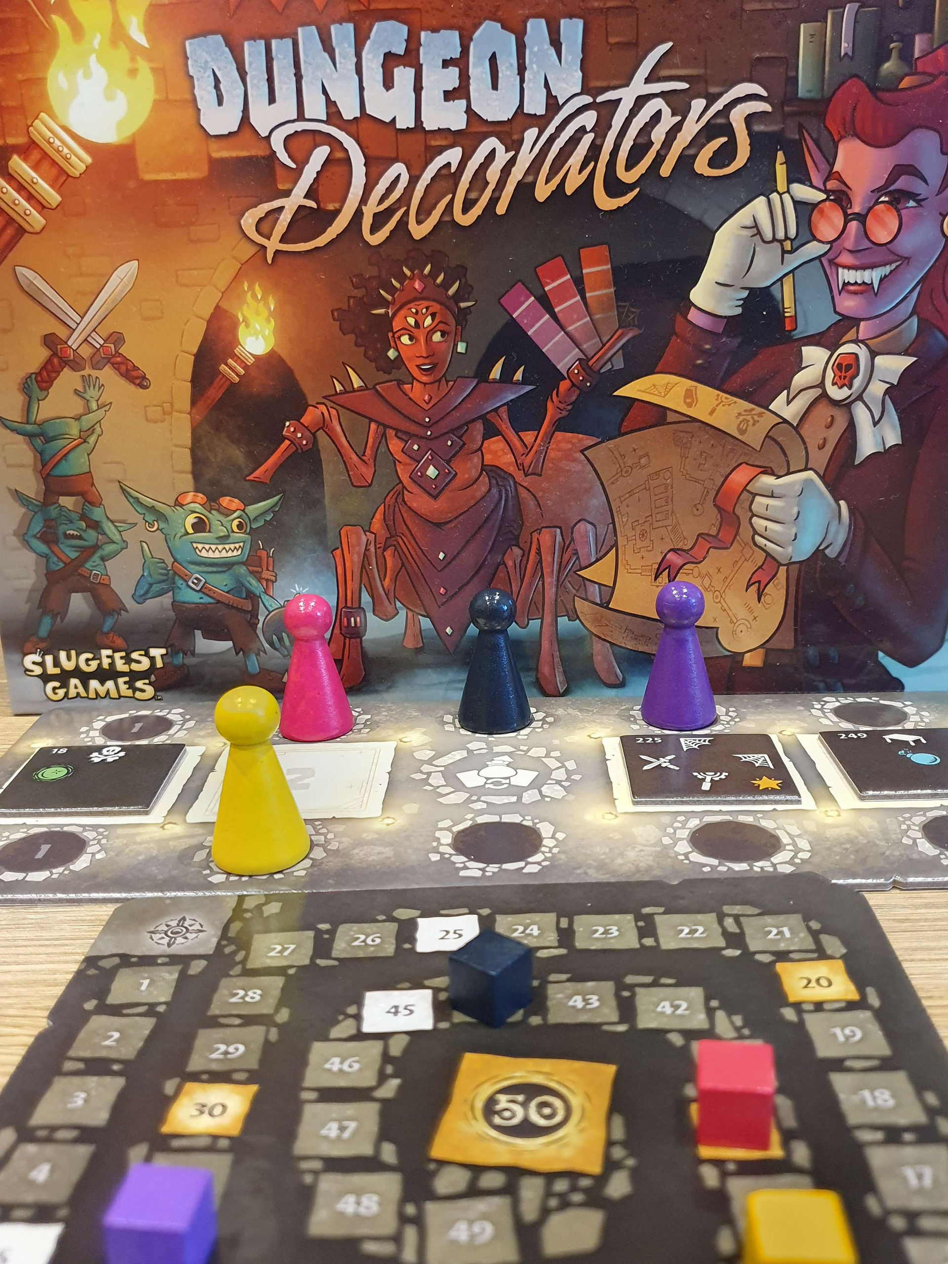 Player pawns from the game Dungeon Decorators- yellow, pink, black and purple. Part of the score tracker can be seen in the foreground 
