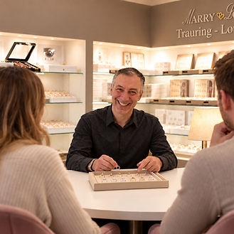 Marry Lou Berater zeigt einem Paar Trauringe am Beratungstisch in der Trauring-Lounge