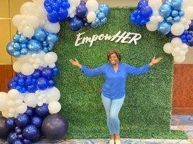 Balloon arch in white and shades of blue along the top and side of a green wall and a sign that says "EmpowerHer"
