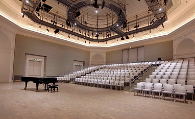 This image shows an empty concert hall, decorated in beige and white, with a grand piano on the floor.