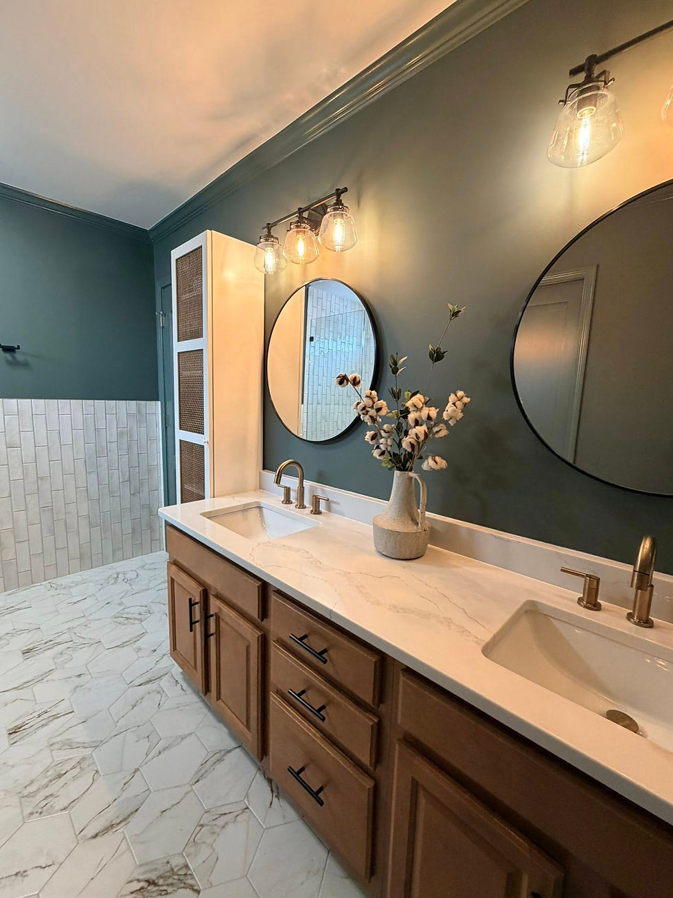 Bathroom with dual sinks, round mirrors, and modern lamps. Green wall, wood cabinets, marble countertop, tile floor, vase with flowers. Cozy vibe in home renovation in Davidson, NC.