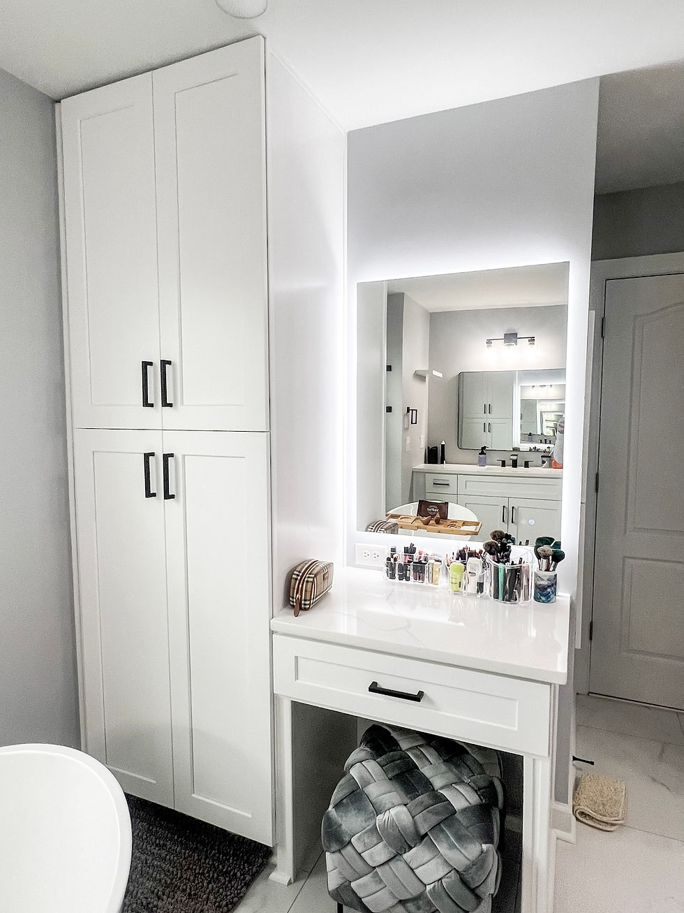 White bathroom with a glowing vanity mirror, makeup display, and a cushioned stool. Minimalist cabinets and a soft rug enhance the clean look.