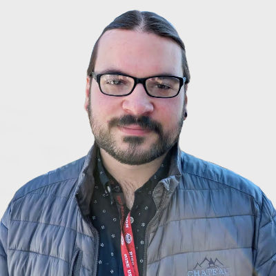 Man with glasses in a gray quilted jacket and patterned shirt, wearing a red lanyard. Neutral expression, plain white background.