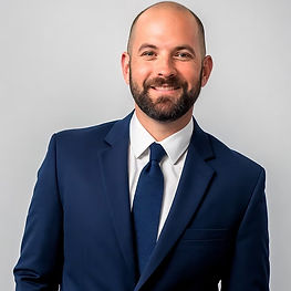 Man in a blue suit and tie smiling against a plain light gray background. Professional and confident demeanor.