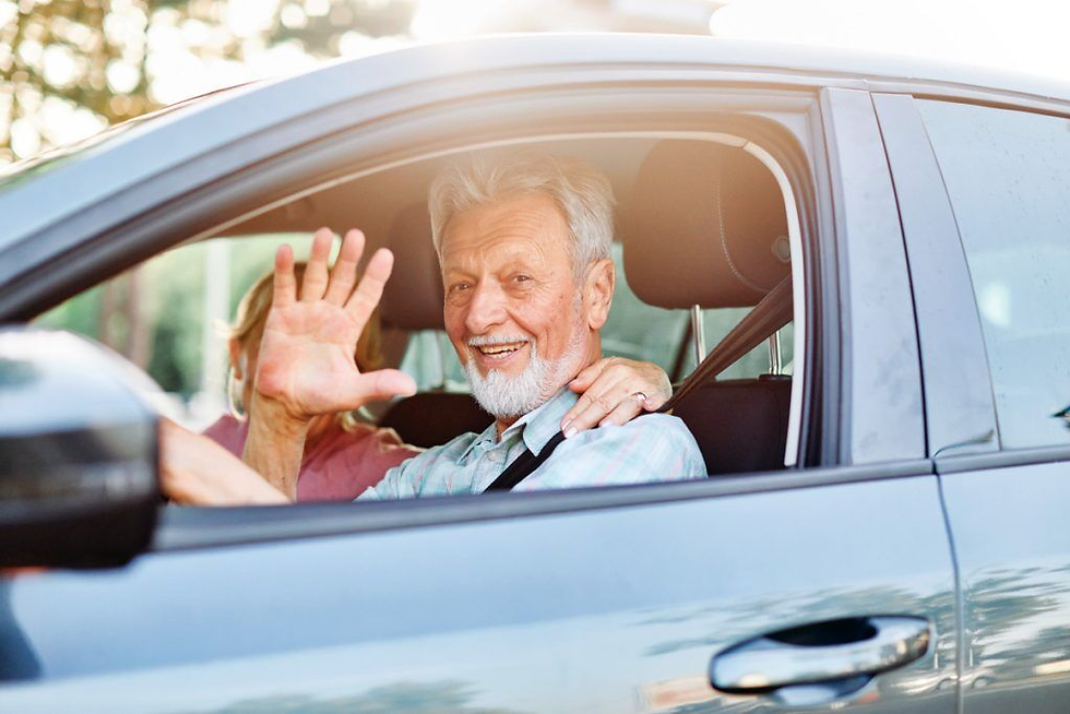 Smiling elderly man waves from a car window, wearing a light blue shirt. Sunlight filters through trees in the background, creating a warm mood.