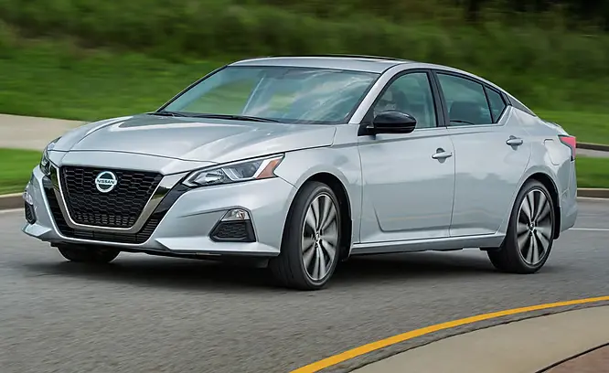 Silver Nissan driving on a curved road with green grass in the background. Sleek design, alloy wheels, and a visible Nissan logo.