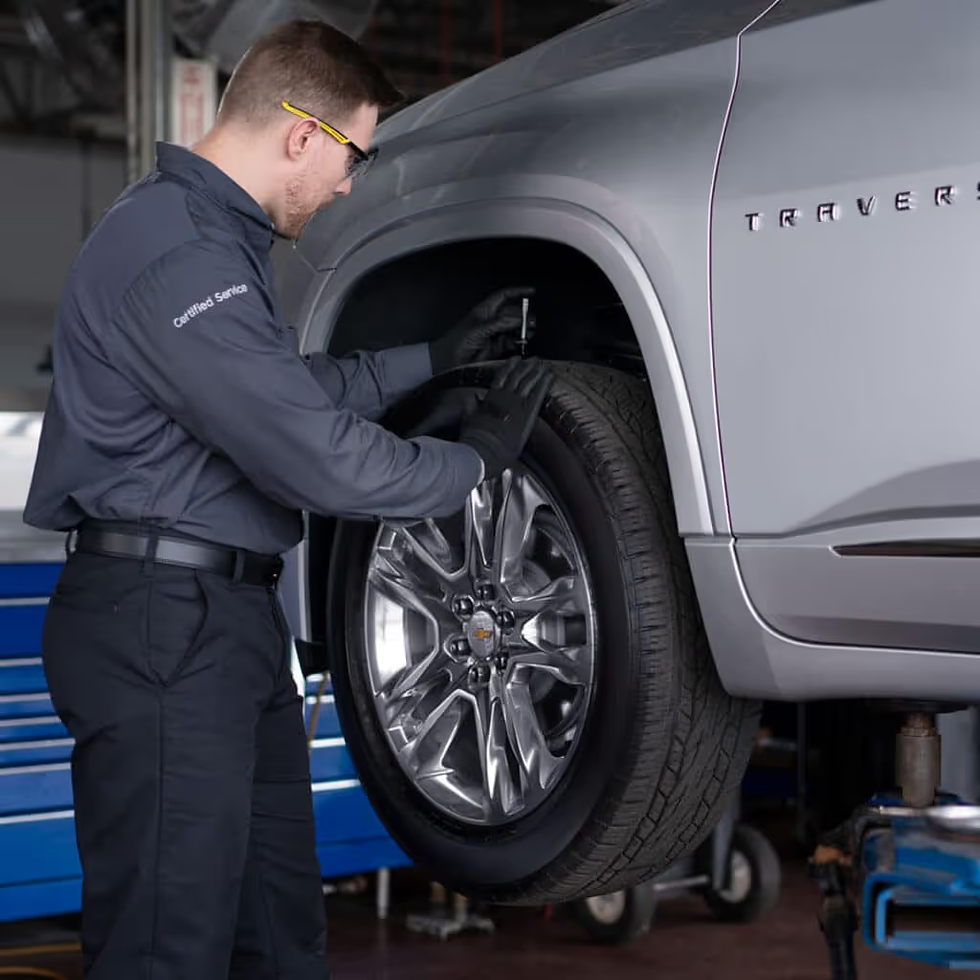 Mechanic in gray uniform adjusts a gray SUV's tire in a garage. Car logo visible. Focused atmosphere with blue equipment nearby.