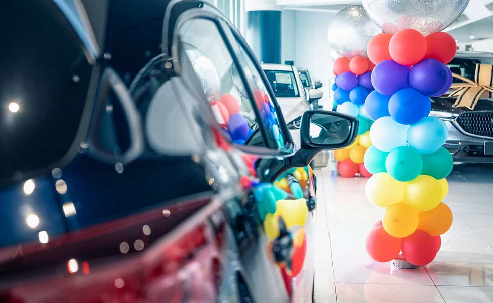 Car showroom with colorful balloon columns reflecting on a shiny car. Bright lighting creates a festive atmosphere.
