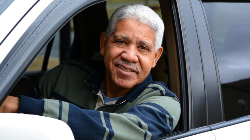 Smiling man in striped sweater sitting in a car, arm resting on window. Background shows a blurred outdoor scene. Mood is cheerful.