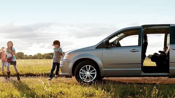 Children run joyfully on grass near an open silver minivan under a clear sky. A sense of freedom and playfulness in a sunny, rural setting.