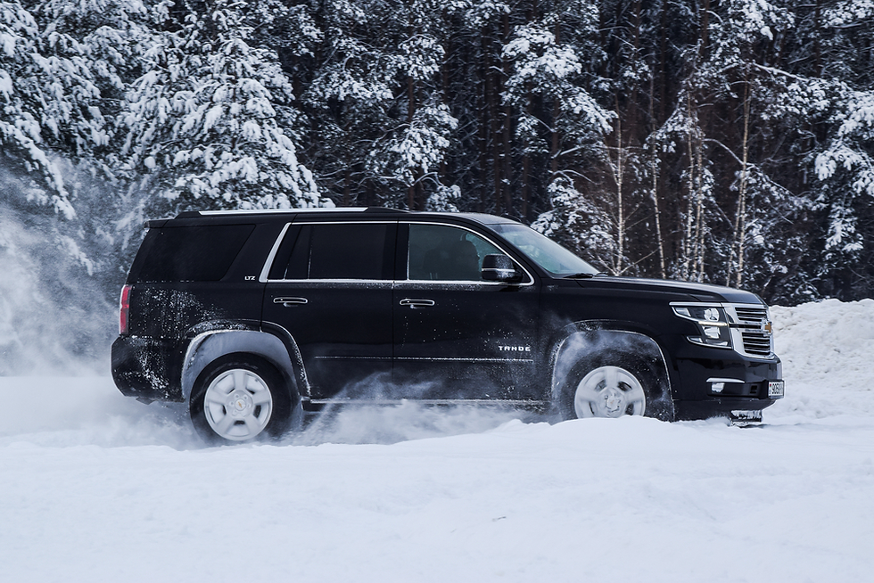Black SUV driving through snowy forest, creating a spray of snow. Winter trees in the background. Vehicle has "LTZ" and "Tahoe" labels.