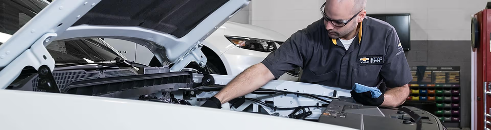 Mechanic inspects engine of a white car in a garage. He wears safety glasses and a Chevrolet uniform. Tools and cars are visible in the background.