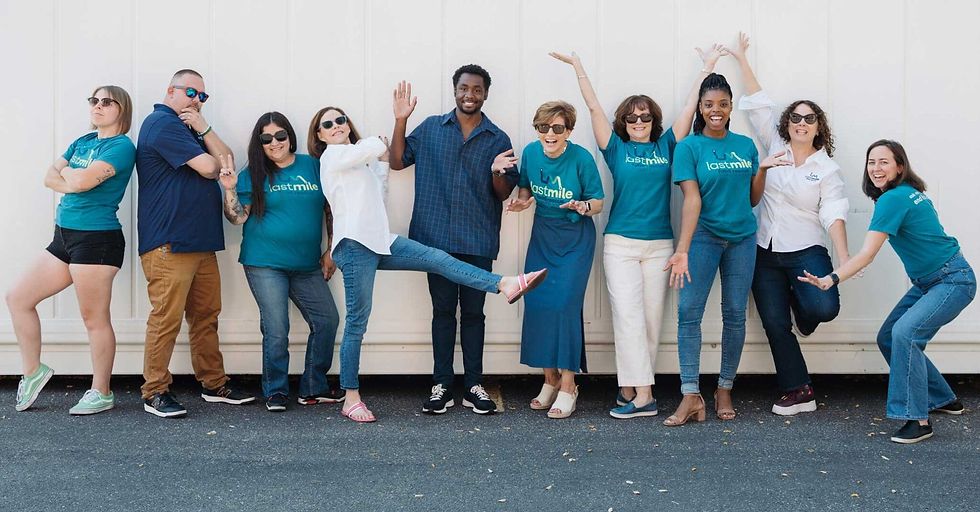 Nine people posing playfully in front of a white wall, wearing "last mile" teal shirts or casual outfits, creating a joyful group vibe.