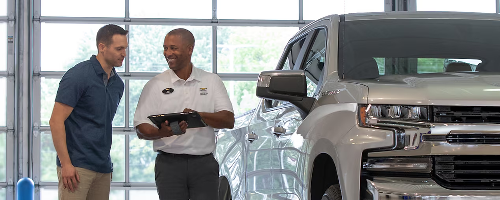 Two men smile and discuss beside a silver truck in a garage. One holds a tablet. Bright light streams through large windows.
