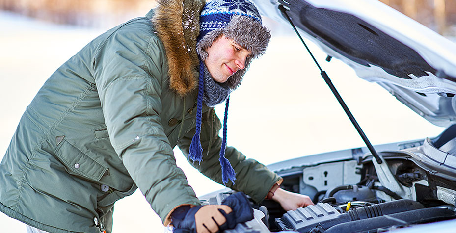 Man in a green parka and blue hat inspects a car engine on a snowy day, with open hood. Winter scene, focused and calm mood.