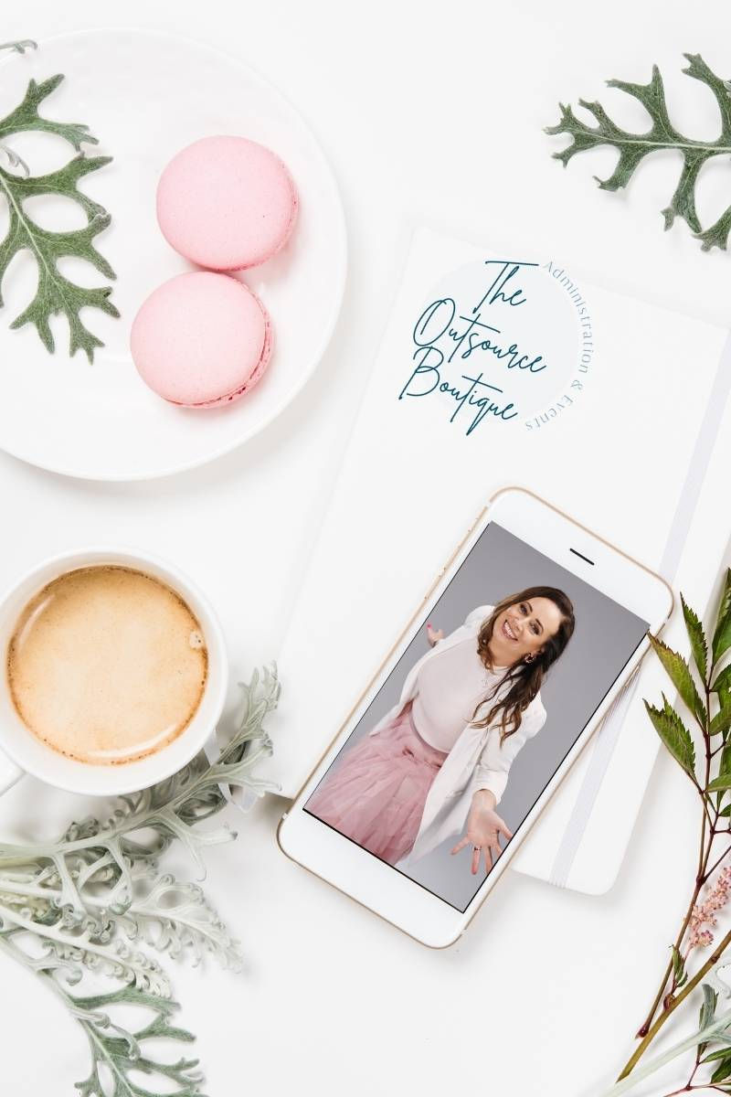 a white saucer with 2 pink macrons beside a white espresso coffee, a white notebook with the words The Outsource Boutique stacked vertically teal, above white iphone with a picture of the founder of the Outsource boutique, Christina Hull, a white lady with long medium brown hair, a white jacket and a pink tule skirt. there are 4 light green plant leaves spaced in the four corners of the picture