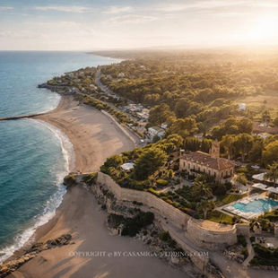 Best wedding venues in Costa Brava by Casamiga Weddings: Aerial view of a coastal villa with a pool, surrounded by lush trees. Adjacent beach and ocean under a golden sunset, creating a serene mood.