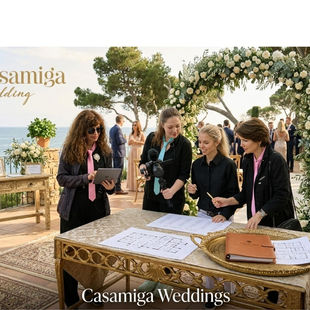 Wedding planner coordinating supplier timing and guest flow for a Costa Brava wedding. Wedding planners check a guest list at a seaside Casamiga Wedding setup with white floral arch, gold tables, and staff in black suits. Foto: Personas/vestuario mejorados mediante IA.