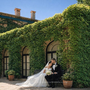 Bride and groom sit on a bench in an ivy-covered courtyard; the bride holds a bouquet, with a soft, romantic mood and watermark text.