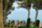 Panoramic sea view from clifftop wedding terrace at Convent de Blanes, Girona — destination wedding Costa Brava 200 guests. Guests mingle on a seaside terrace with wooden decking, surrounded by trees and ocean views. Lively and colorful gathering.
