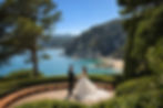 Clifftop sculpted terraces of Jardines de Santa Clotilde with sea view, Lloret de Mar, Costa Brava — wedding ceremony venue 2026 by Casamiga Weddings. Bride and groom hold hands on a terrace overlooking a scenic coastline with lush greenery, clear blue water, and rocky hills under a clear sky.