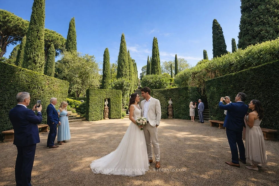 Jardines de Santa Clotilde wedding venue Spain, Costa Brava, Casamiga Weddings. Bride and groom smiling in a lush garden, surrounded by guests taking photos. Tall trees and hedges frame the sunny scene.