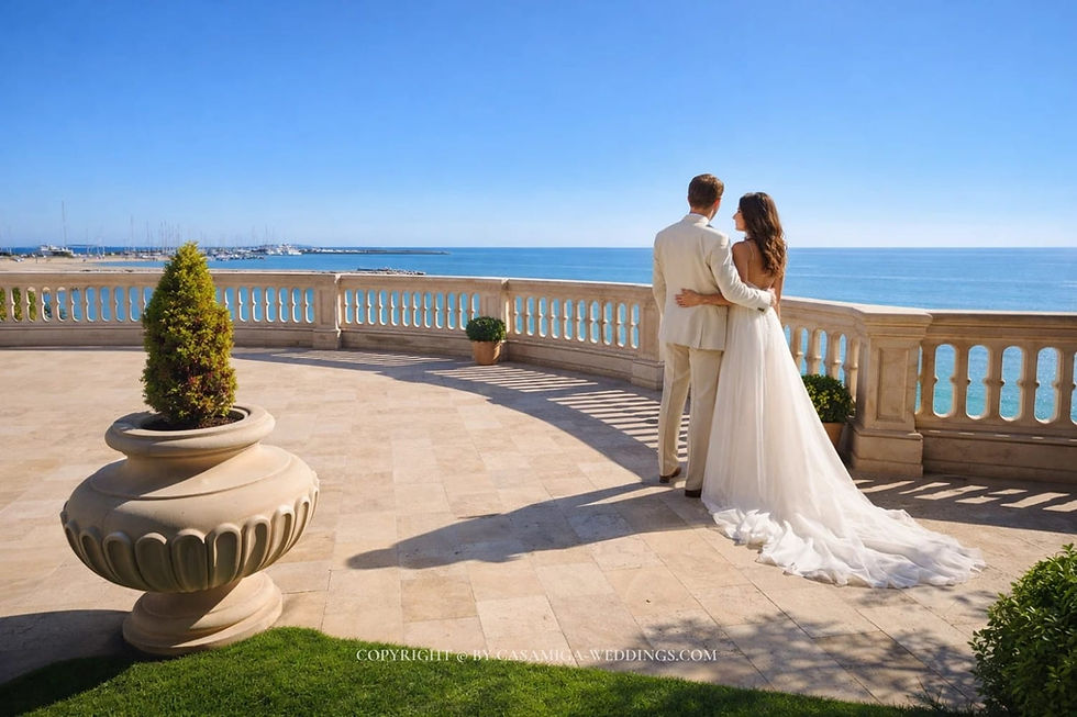 Barcelona destination wedding 2027 skyline ceremony Casamiga Weddings. A couple in wedding attire embraces on a stone terrace overlooking the sea. It’s a sunny day with a blue sky. Potted plants decorate the space.